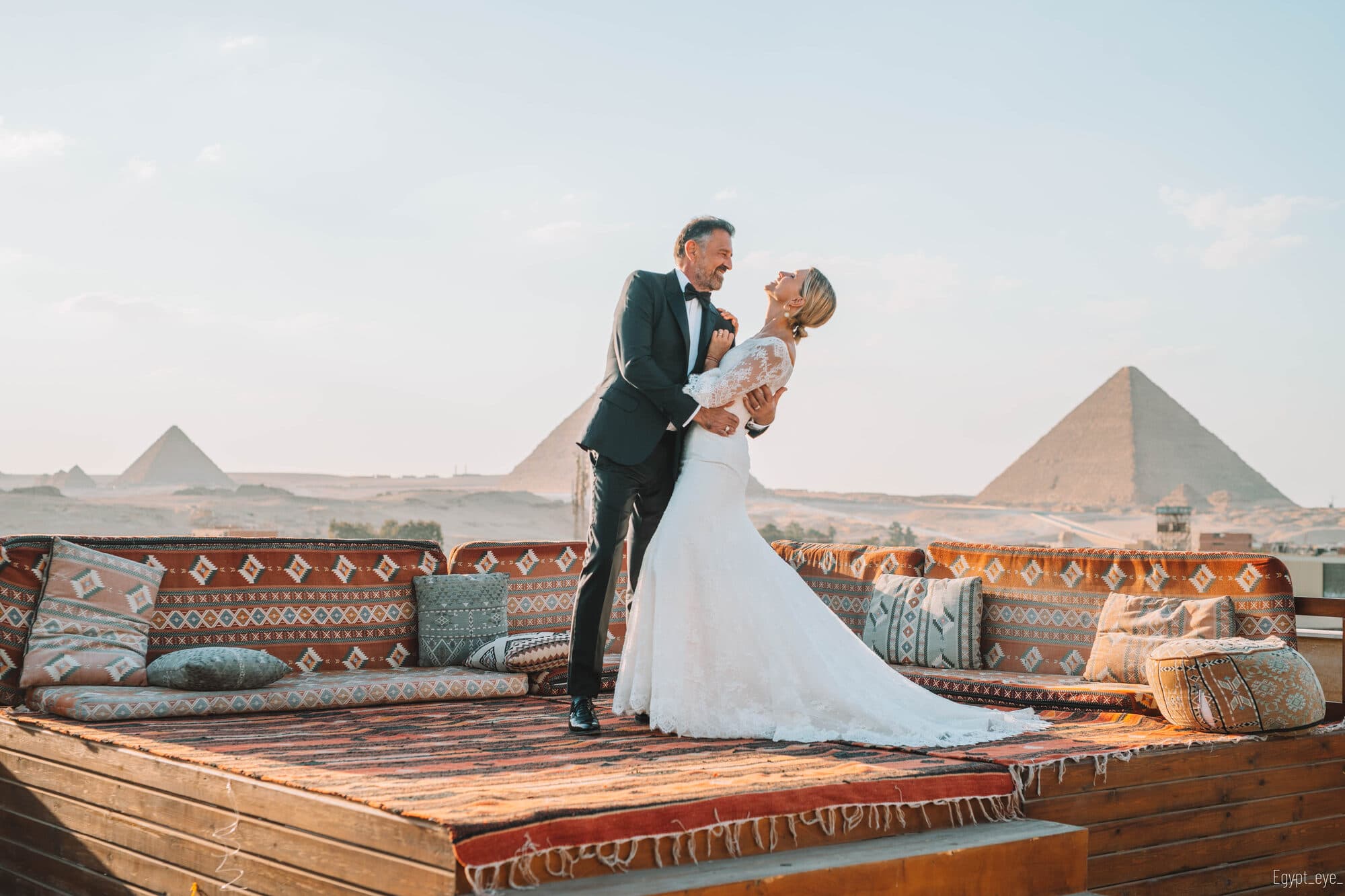 A couple on the Giza rooftop at sunrise, he in black tie with his arm around her waist, she laughing in a long lace gown, three pyramids across the horizon behind them