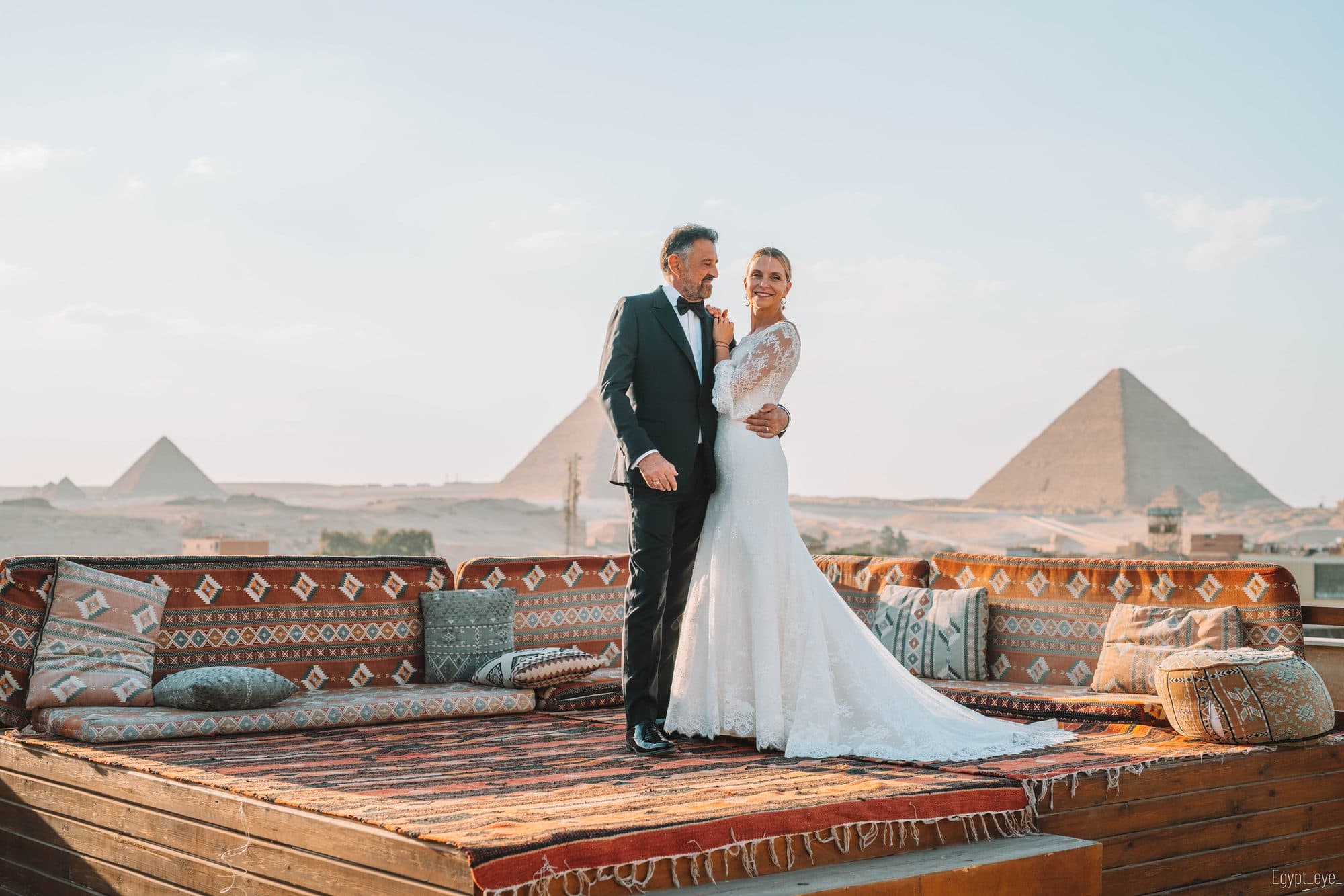 A couple in formal dress on the Giza rooftop at golden hour, her train laid out behind her, two pyramids clear on the horizon