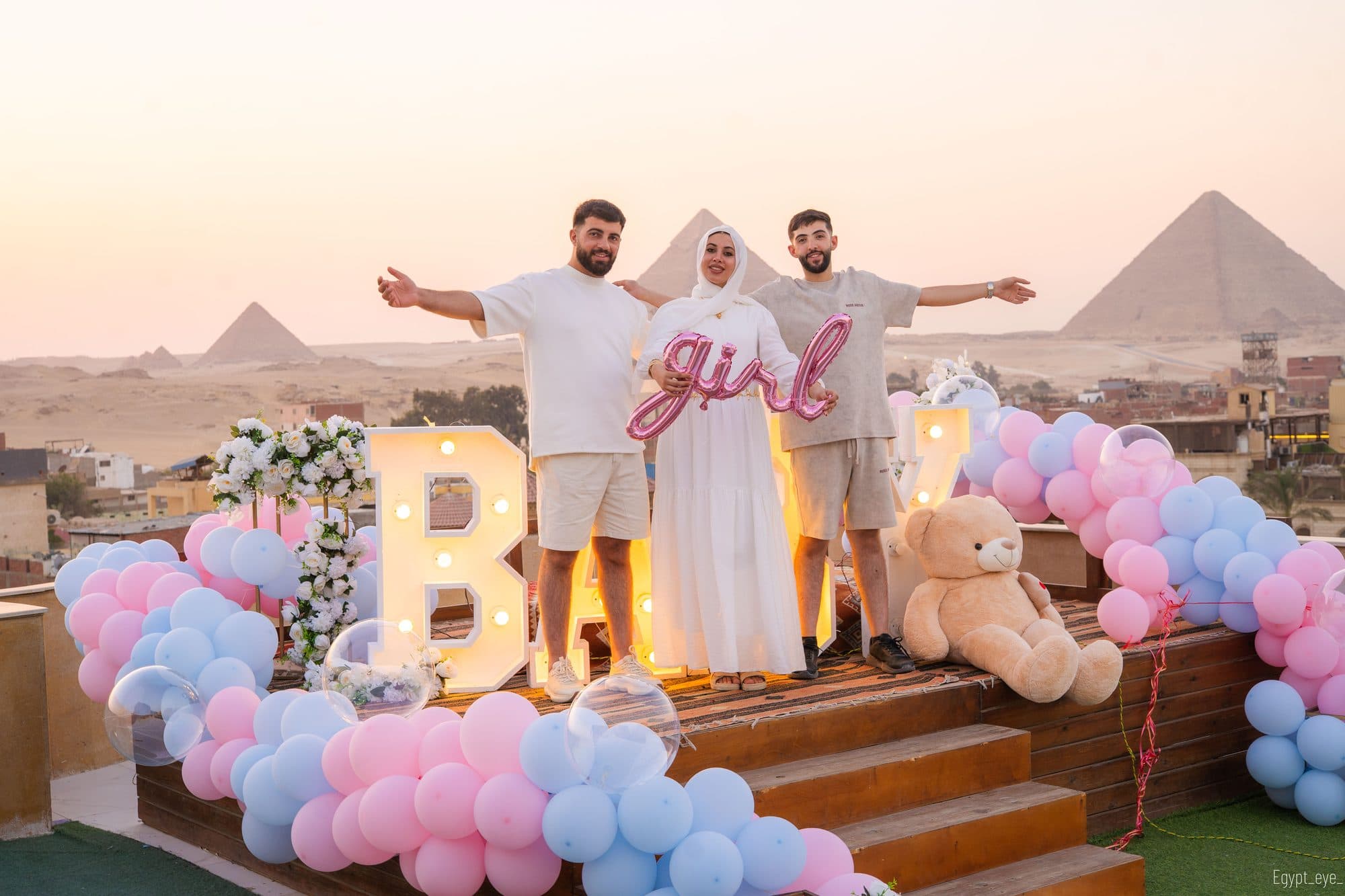 Parents and a guest with arms thrown wide beside lit BABY letters and a pink and blue balloon arch, the pyramids behind them at sunset