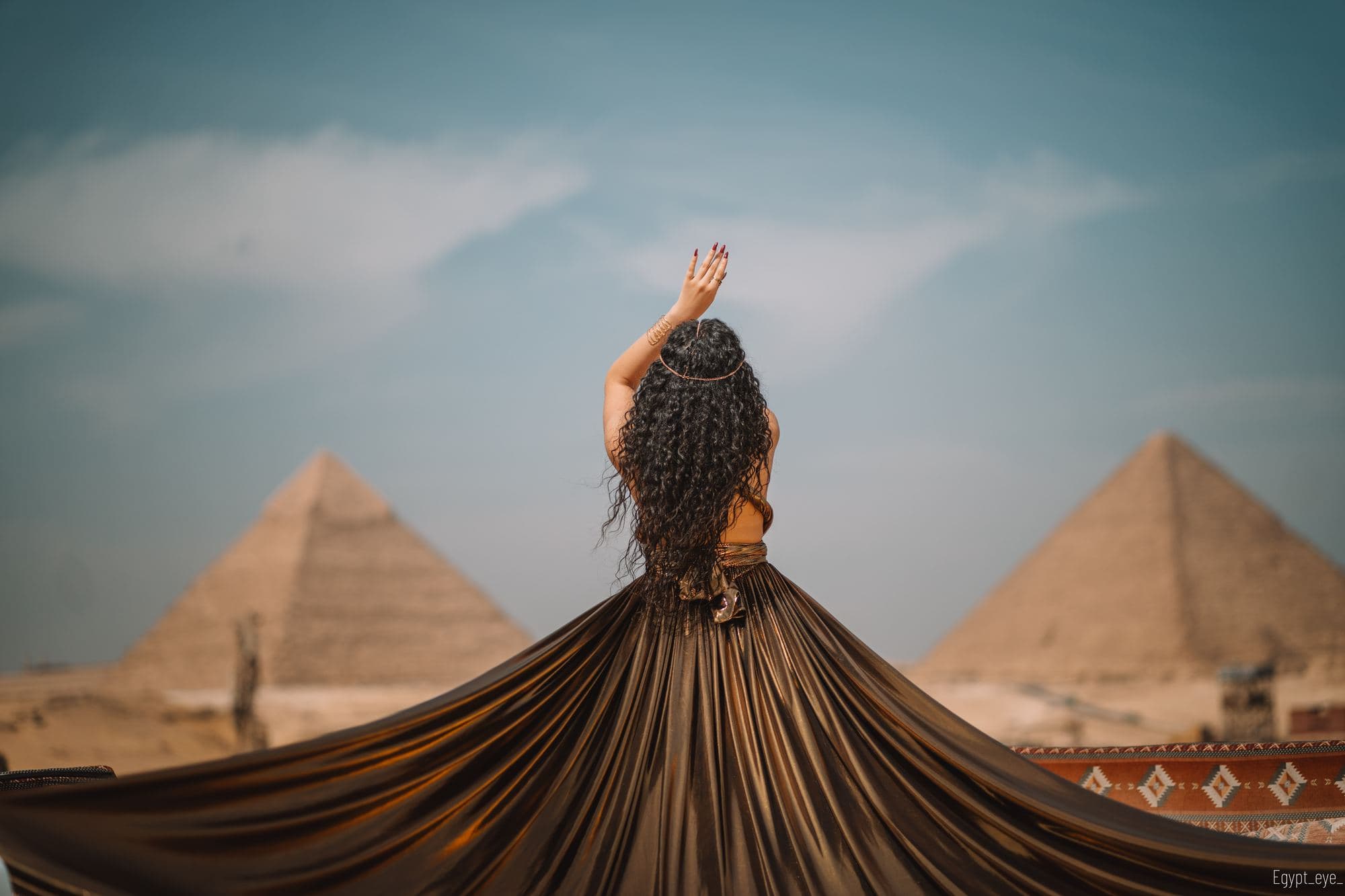 A gold dress fanned out across the Giza rooftop, the wearer facing the two pyramids
