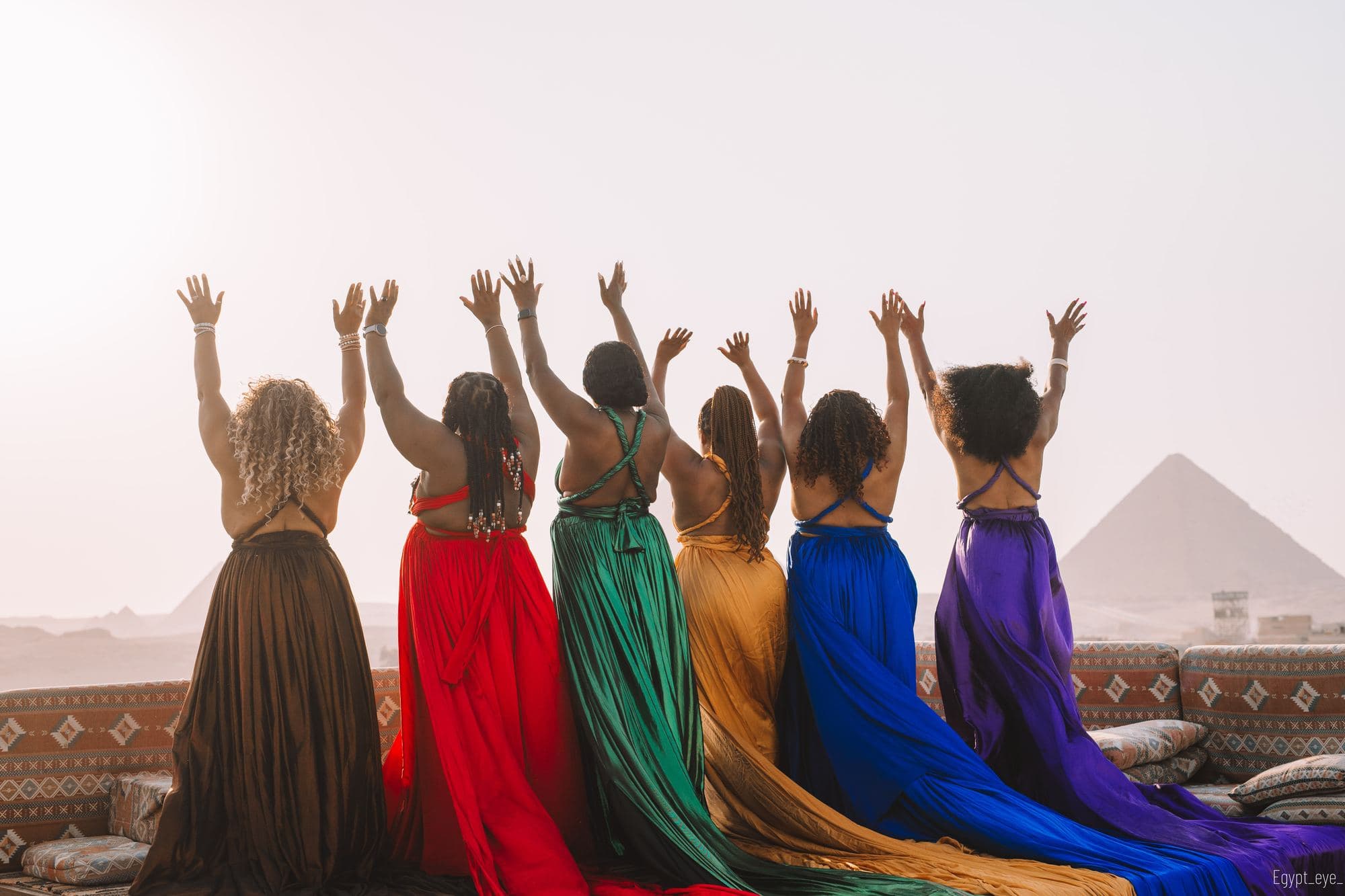 Six women on the Giza rooftop with their backs to the camera, arms raised, each in a different colour, a pyramid on the skyline