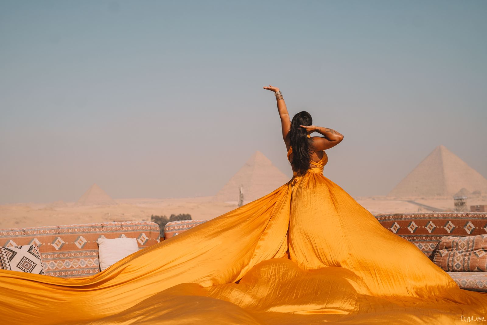 The Ginger dress from behind, amber silk fanned wide across the rooftop under the pyramids