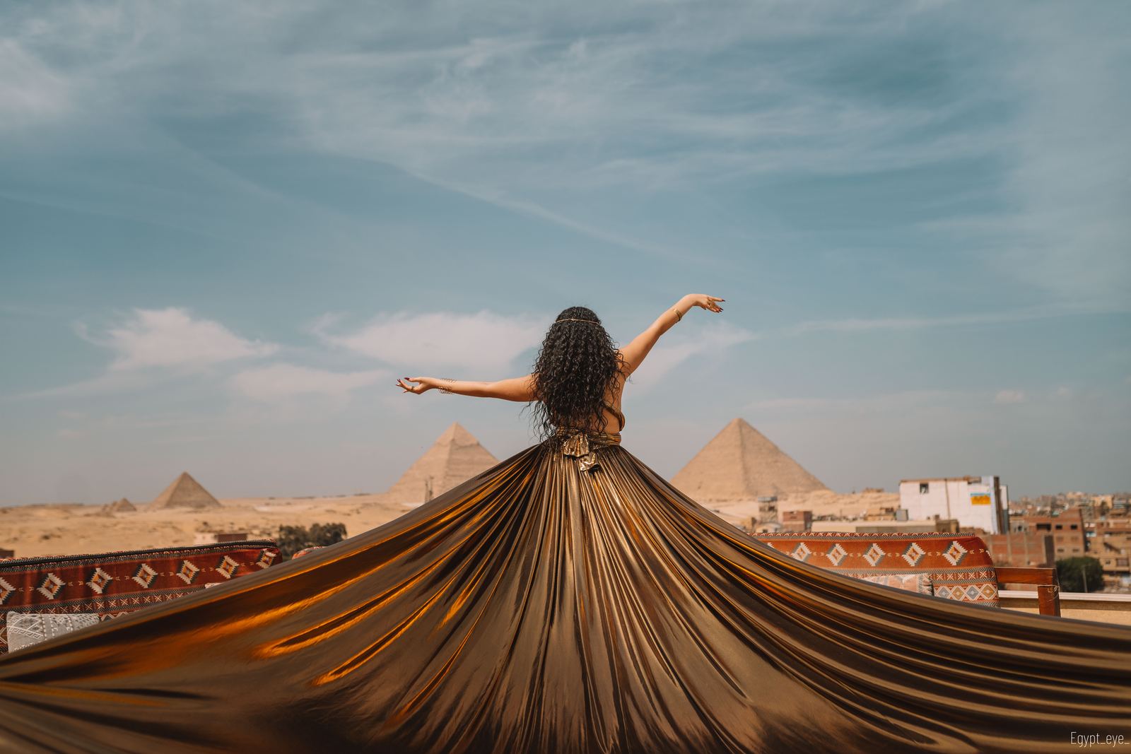 The Golden dress opened into a full fan, arms outstretched, the pyramids behind the rooftop